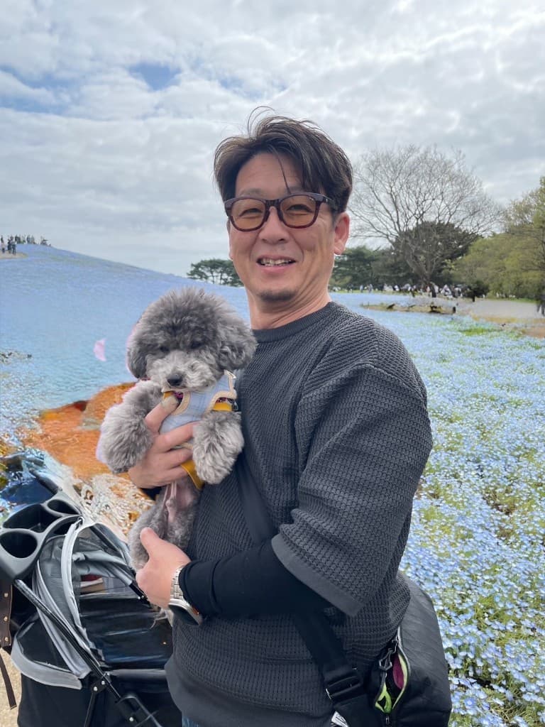 代表取締役 金子 誠博が屋外の花畑の前で小型犬を抱いている様子